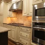 Current image: Cream kitchen with a gas cooktop, stainless range hood, and built-in double ovens set among light cabinets and a warm backsplash.